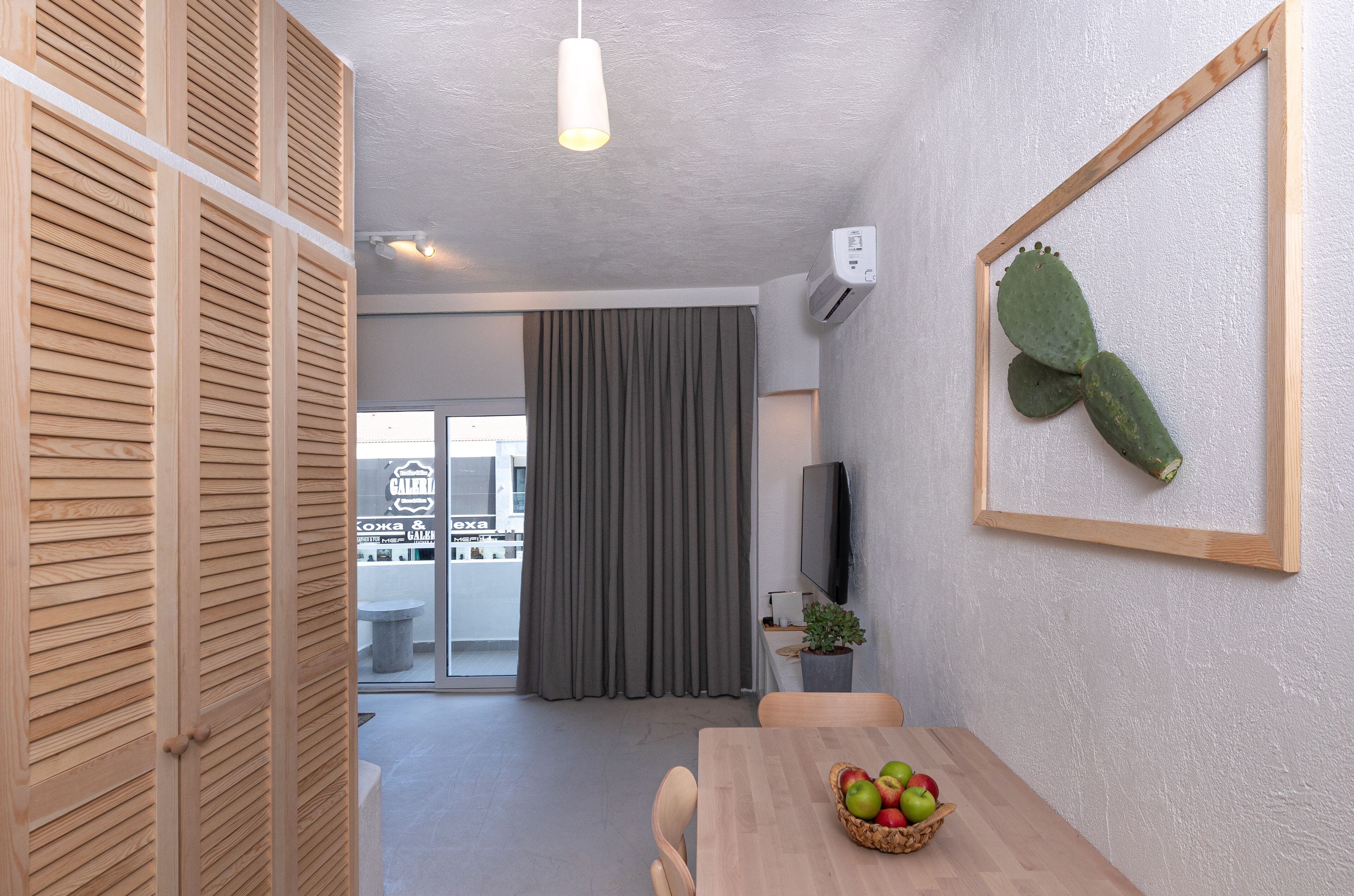 A modern room with a wooden door, a dining table with a fruit bowl, a cactus decoration, and a television on the wall.