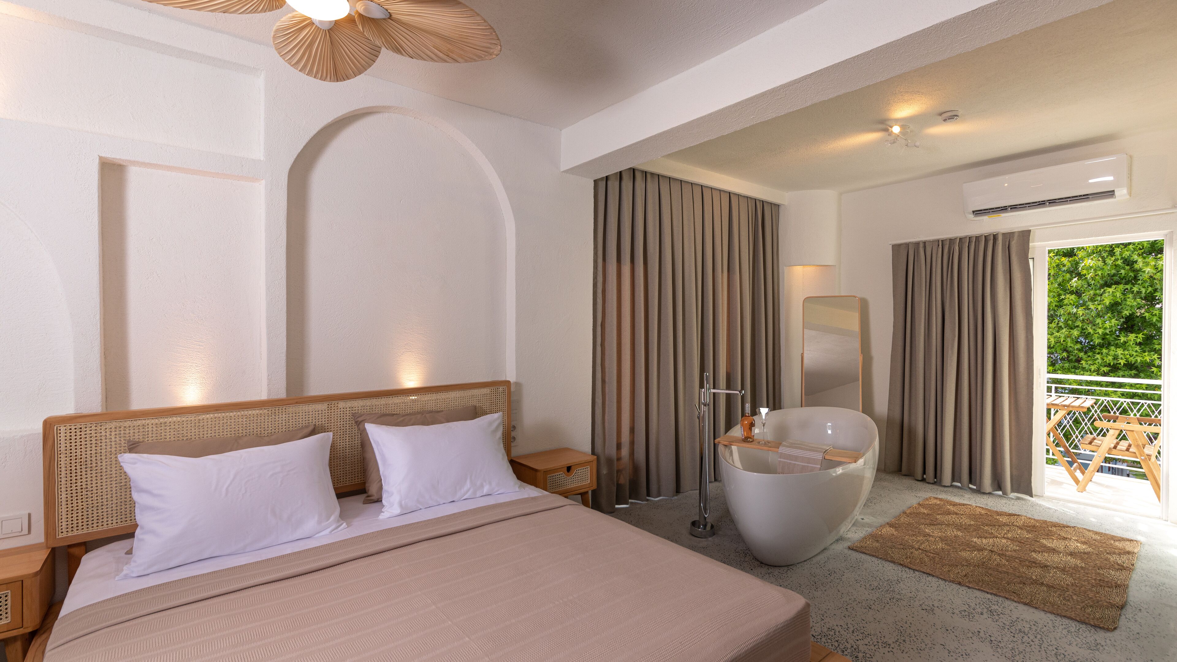 A modern hotel room with a large bed, a bathtub, a mirror, and a balcony with a view of greenery.