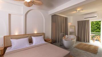 A modern hotel room with a large bed, a bathtub, a mirror, and a balcony with a view of greenery.