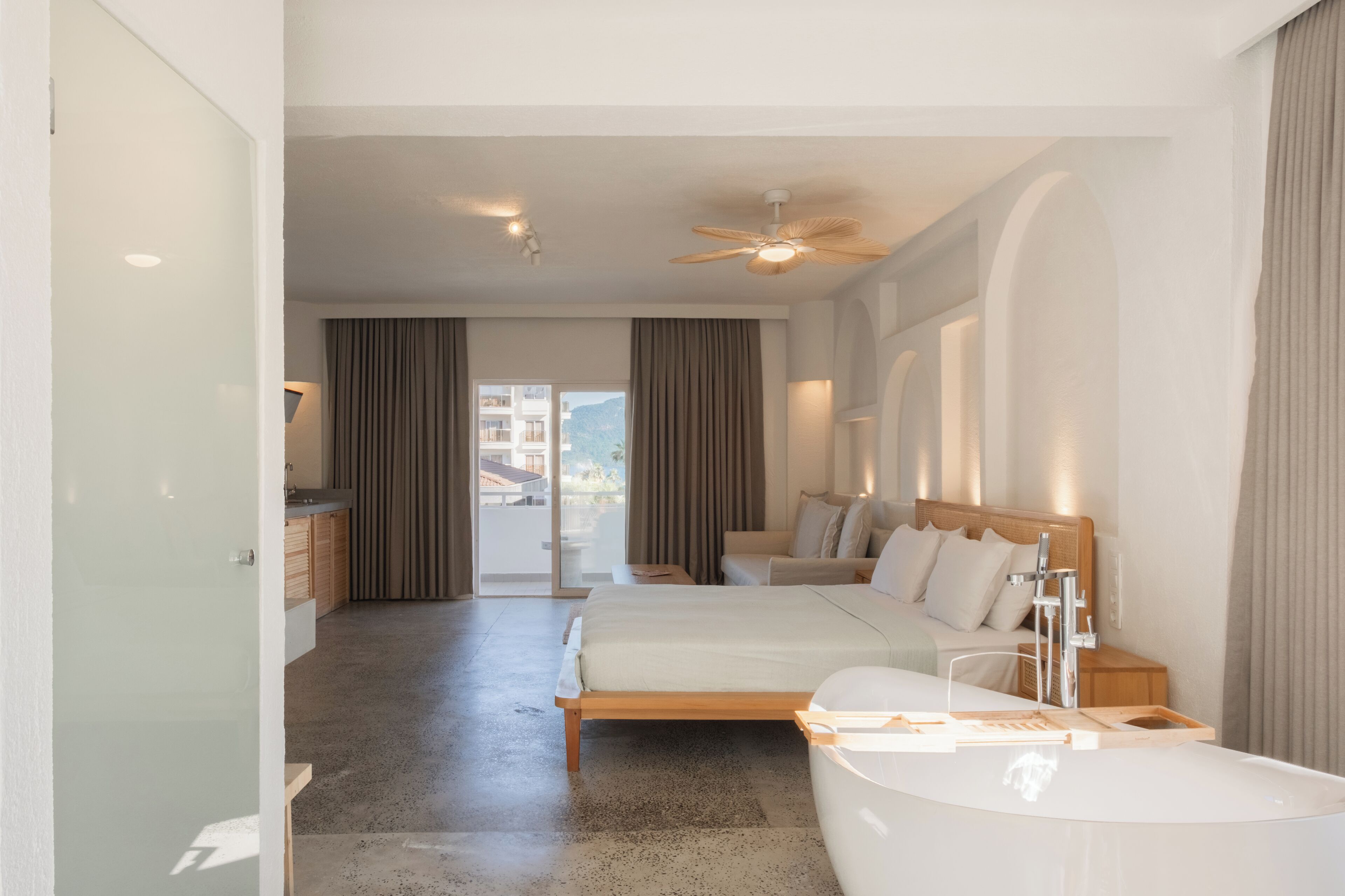 A modern hotel room with a large bed, a separate sitting area, a bathtub, and a view of the mountains.