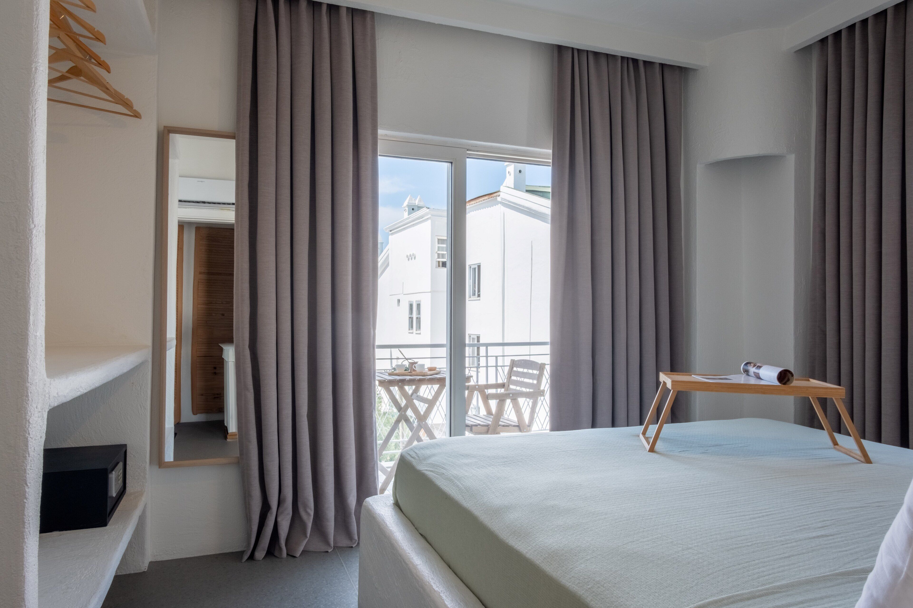 A hotel room with a bed, a wooden table, a chair, a balcony with a table and chairs, and a large window with curtains.