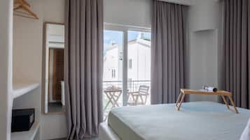 A hotel room with a bed, a wooden table, a chair, a balcony with a table and chairs, and a large window with curtains.