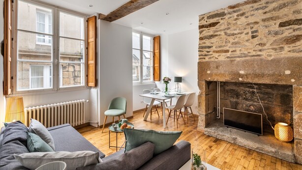 A cozy living room with a stone fireplace, a dining area with a white table and chairs, and a large window with wooden shutters. - Le Cerf Volant - une pépite à l'Intramuros (Saint-Malo)