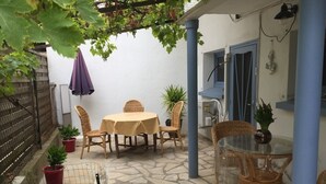 An outdoor patio with a round table and chairs, a glass-top table, potted plants, and a pergola with vines. - Appartement Rez de Jardin Piscine Chauffée, Barbecue, Plancha (Argelès-sur-Mer)