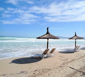 Ein Strand mit zwei Reetsonnenschirmen und weißen Liegestühlen.