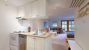 Cocina compacta con armarios blancos, estufa y lavamanos. Hay una zona de comedor con sofá y cama al fondo.