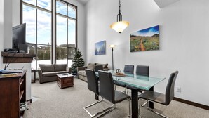 A modern living room with a glass dining table, black chairs, a fireplace, a TV, and large windows with a view of a snowy landscape.