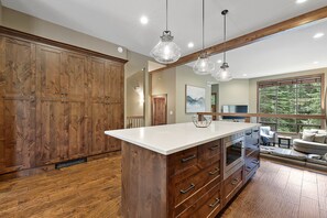 A spacious kitchen with wooden cabinets, a large island with drawers, pendant lights, and a view of a living area with a sofa and a window overlooking a forest.