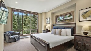 Ein Schlafzimmer mit einem großen Bett, einem Sessel, einem Nachttisch, einer Kommode und einem Fenster mit Blick auf Bäume.