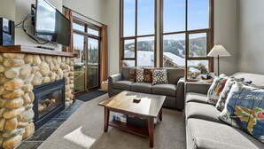 A living room with a stone fireplace, a large window with a view of a snowy landscape, a gray sofa, and a coffee table. - Pet Friendly, Ski In/Ski Out, 3 Bedroom Condo in Stone's Throw! (Sun Peaks)