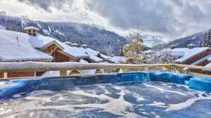 A hot tub area with a wooden deck and a view of snow-covered buildings and mountains. - Pet Friendly, Ski In/Ski Out, 3 Bedroom Condo in Stone's Throw! (Sun Peaks)