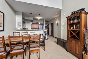 A dining area with a wooden table and chairs, a kitchen in the background, and a wooden cabinet with glassware.