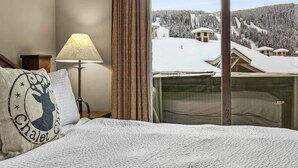 A hotel room with a bed, a lamp, a window with a view of a snowy mountain landscape. - Pet Friendly, Ski In/Ski Out, 3 Bedroom Condo in Stone's Throw! (Sun Peaks)