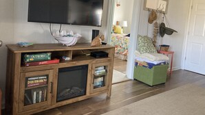 A living room with a TV mounted on the wall, a wooden entertainment center with books and board games, a green cushioned bench, and a door leading to another room. - Just steps to the beach, bar, and playground park! (Surf City)