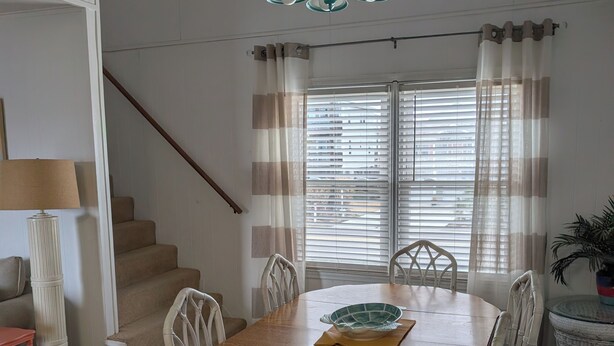 A dining area with a wooden table, white wicker chairs, a hanging light fixture, and a staircase. - Just steps to the beach, bar, and playground park! (Surf City)