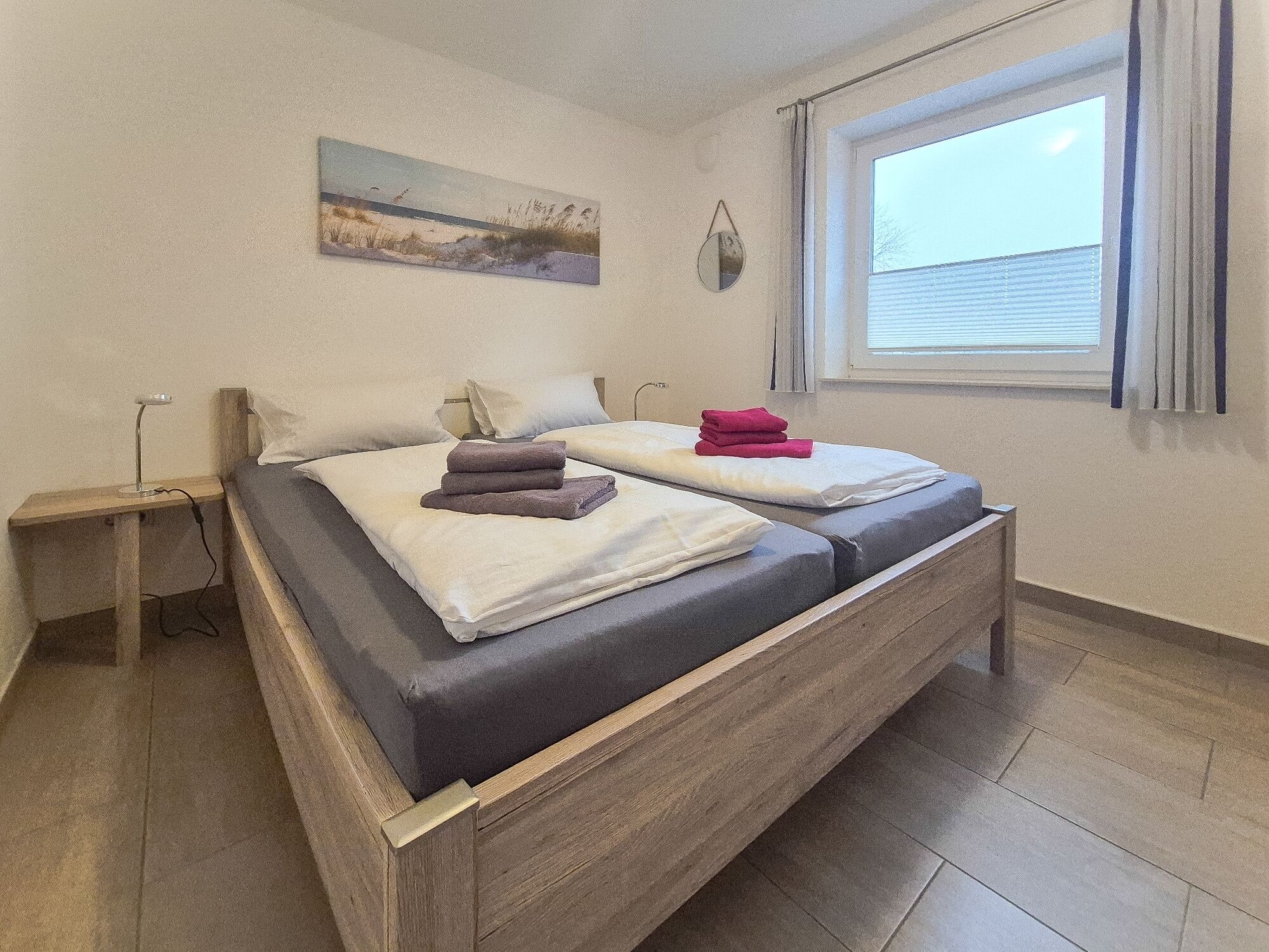 A double bed with a wooden frame, a bedside table, a window with blinds, and a painting on the wall.