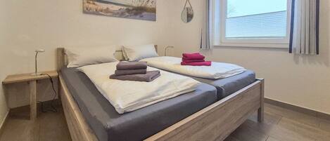 A double bed with a wooden frame, a bedside table, a window with blinds, and a painting on the wall.