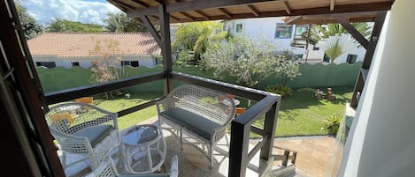 A covered outdoor seating area with wicker chairs and a table, overlooking a well-maintained garden and neighboring buildings.