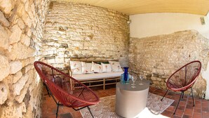 Una habitación de paredes de piedra con un sofá y dos sillones rojos, una mesa redonda con un florero y vasos, y una alfombra estampada.