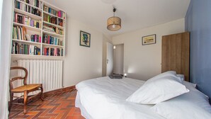 Un dormitorio con una amplia estantería, una cama con ropa de cama blanca, una silla de madera y un radiador.