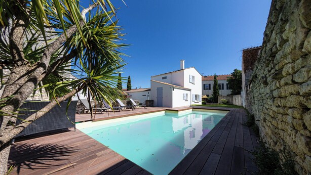 Una piscina con cubierta de madera, rodeada por un muro de piedra y un edificio blanco con techo de tejas.