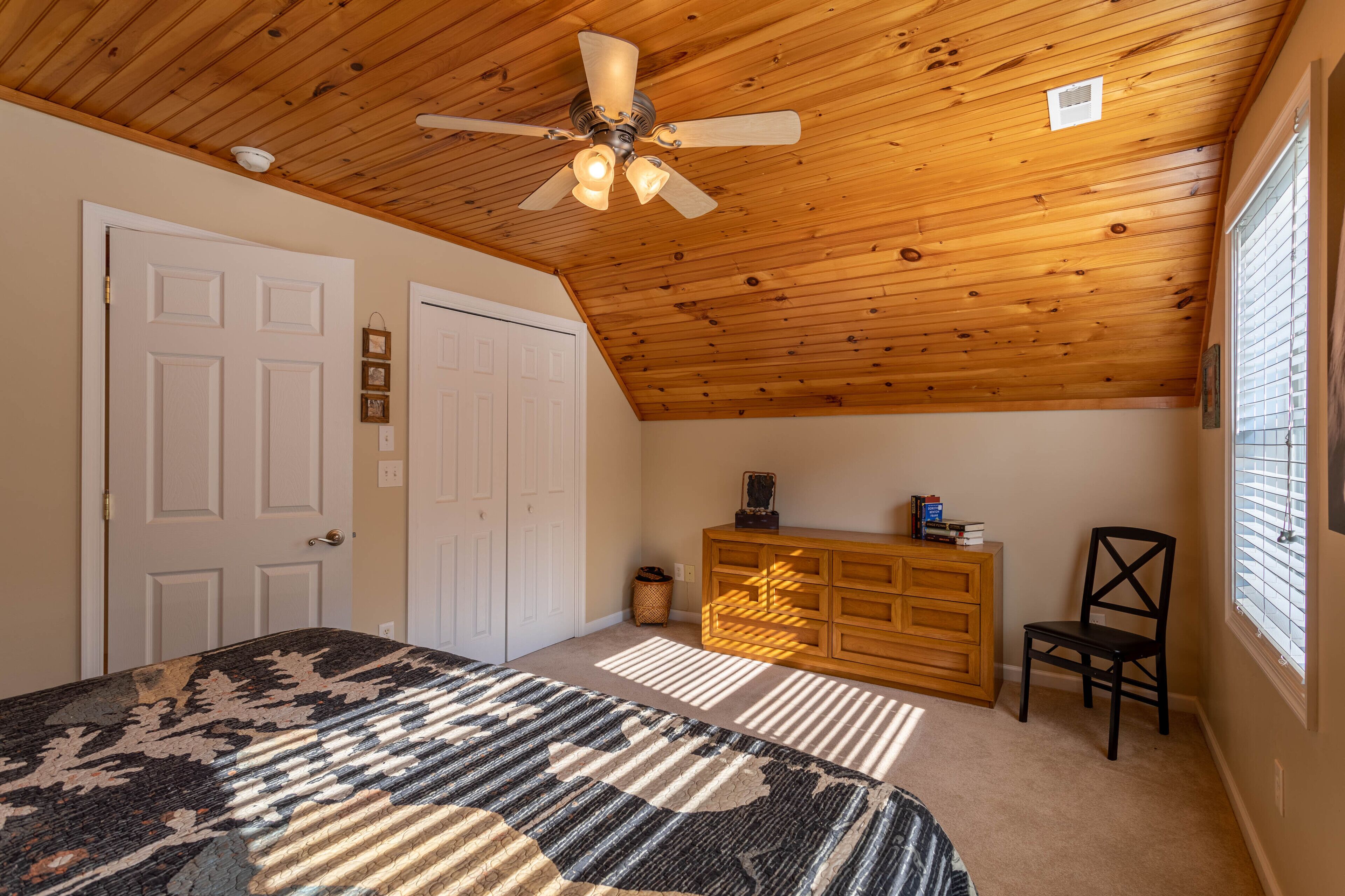 A bedroom with a ceiling fan, a bed with a patterned bedspread, a dresser, a chair, and a window with blinds.