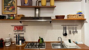 A kitchen with a gas stove, a sink, and various kitchen utensils. There are shelves with baskets and jars, and a framed poster on the wall. - Living Rencio casa vacanza a Bolzano (Bolzano)
