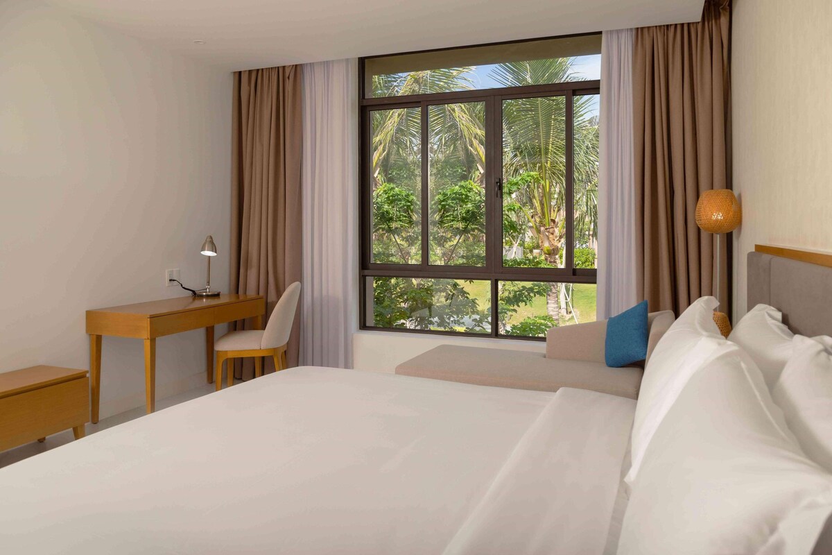 A hotel room with a large bed, a desk with a chair, a lamp, and a view of palm trees through a window.