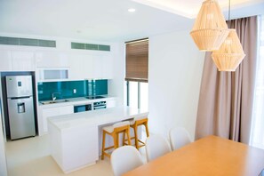 Una cocina moderna con armarios blancos, horno empotrado y refrigerador de acero inoxidable. Hay un comedor con mesa de madera y sillas blancas.