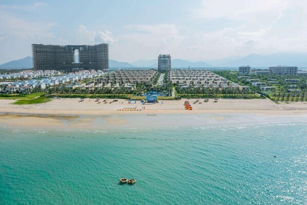 A coastal resort with a sandy beach, a pool, and a large building complex in the background.