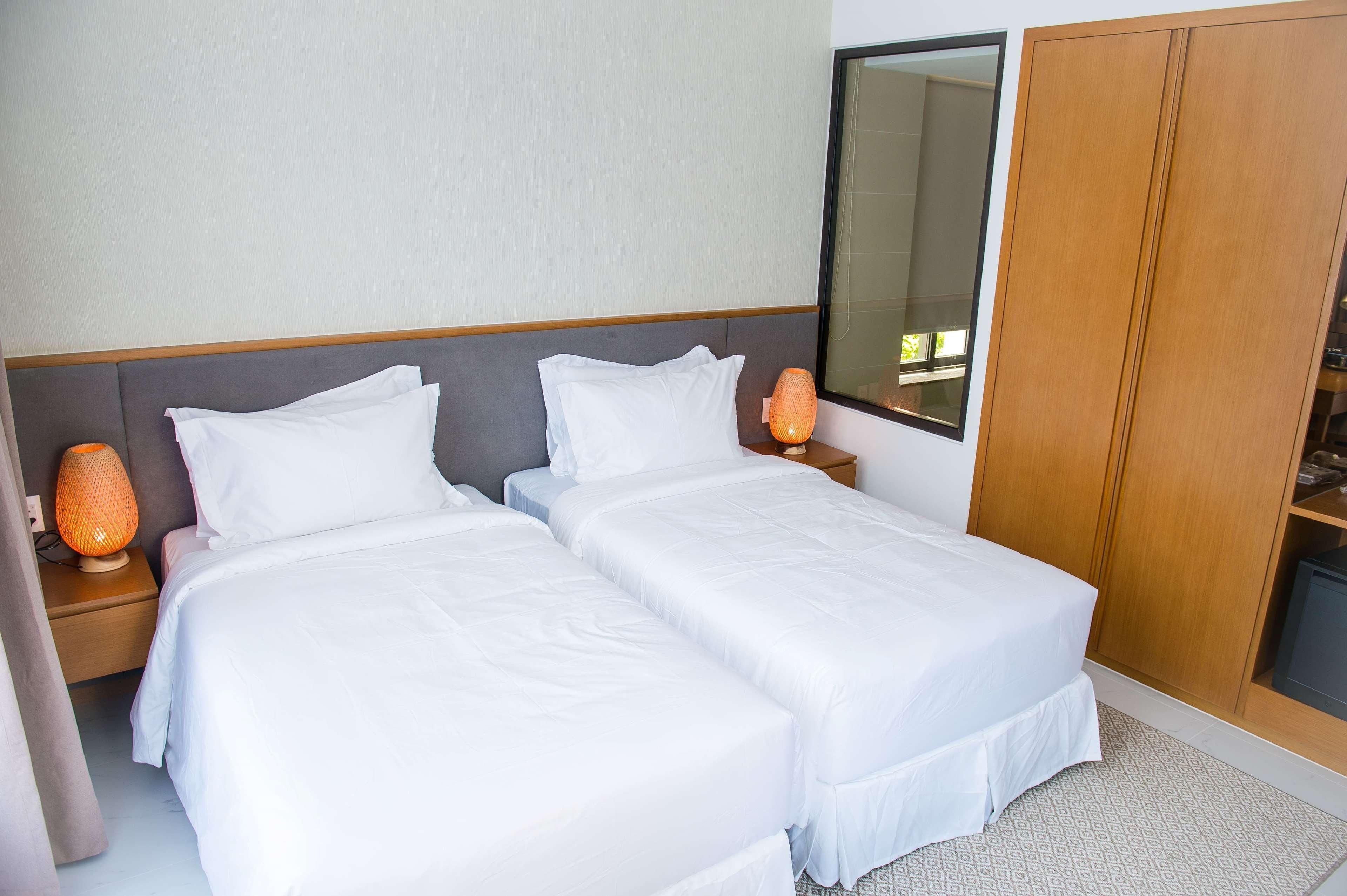A hotel room with two single beds, a wooden wardrobe, a nightstand with a lamp, and a large mirror.