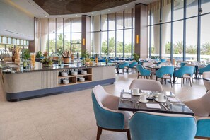 A modern restaurant with a central bar, blue chairs, and large windows offering a view of palm trees. - Wyndham Garden Cam Ranh Resort (Cam Ranh)