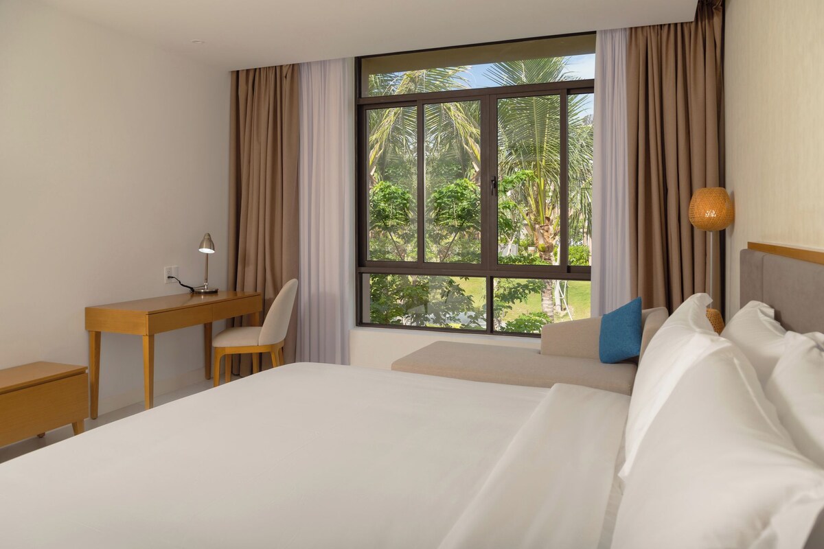A hotel room with a large bed, a desk, a chair, and a window with a view of greenery.