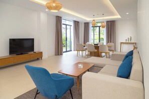 A modern living room with a flat-screen TV, a sofa, a blue chair, a wooden coffee table, and a dining area with chairs and a table. - Wyndham Garden Cam Ranh Resort (Cam Ranh)