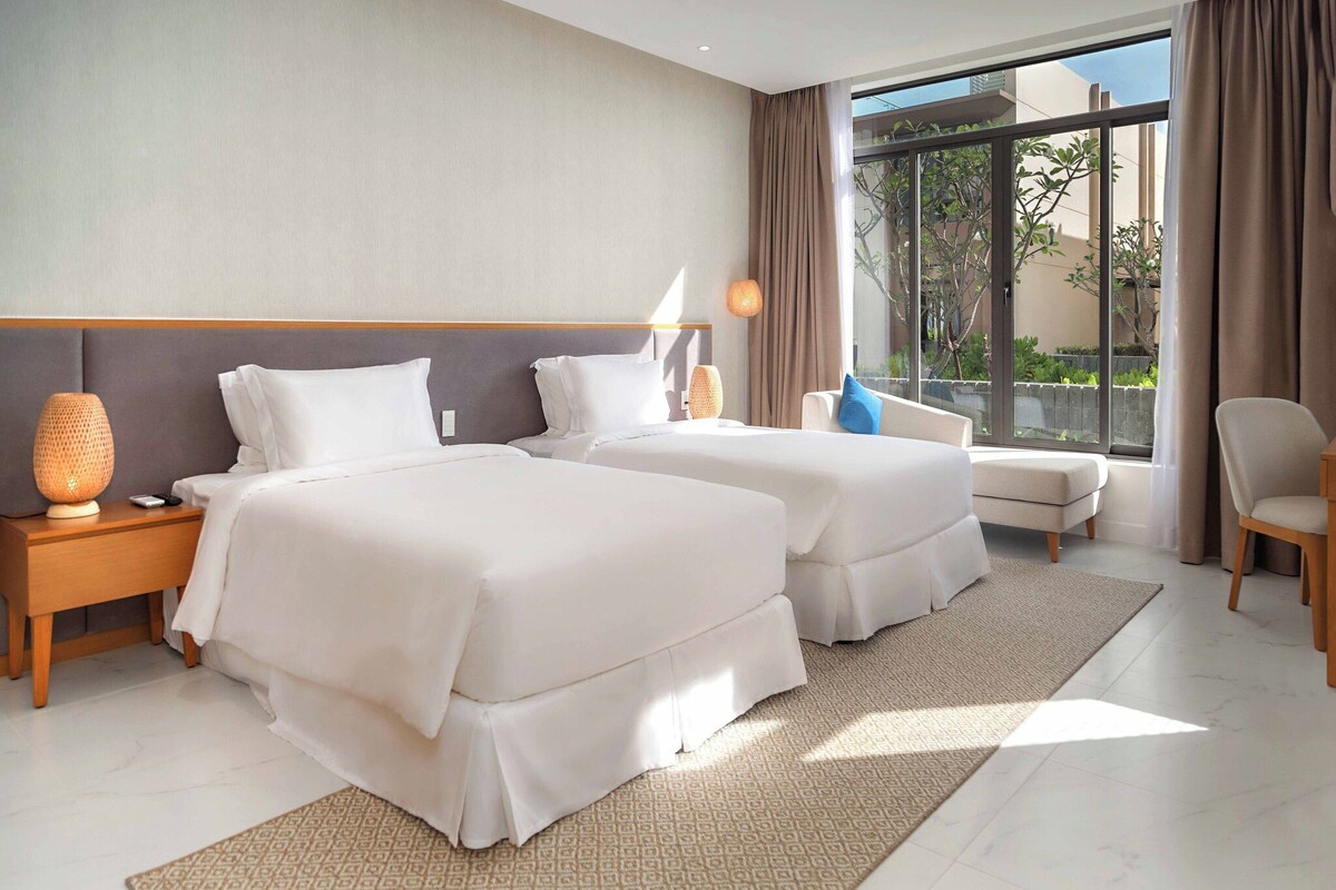 A hotel room with two beds, a sofa, a chair, a lamp, and a view of the outdoors.