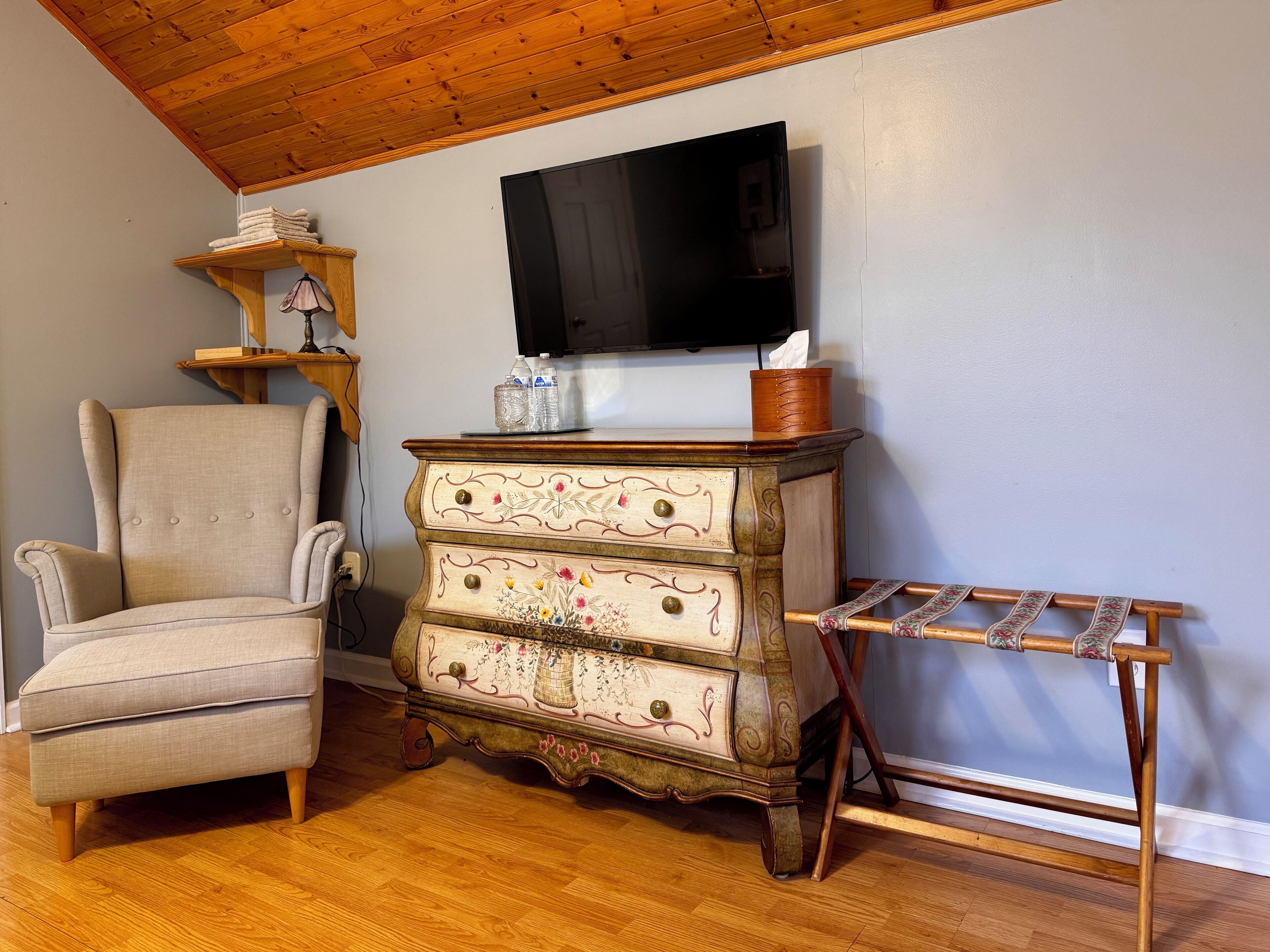 A living room with a wooden ceiling, a beige armchair, a vintage dresser with floral patterns, a wooden drying rack, and a flat-screen TV mounted on the wall.