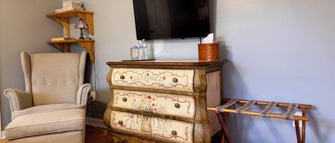 Ein Wohnzimmer mit Holzboden, einem beigefarbenen Sessel, einer Vintage-Kommode mit floralem Muster, einem Wäscheständer aus Holz und einem flachbildfernseher an der Wand.
