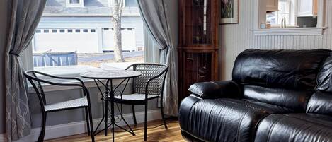 A living room with a black leather sofa, a round table with two chairs, a wooden cabinet, and a view of a house through a window with curtains.