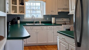 Een keuken met witte kastjes, een groen aanrecht, een roestvrijstalen koelkast en een raam met uitzicht op huizen.
