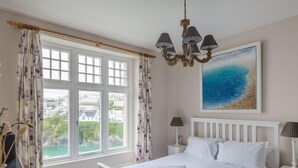 Ein Schlafzimmer mit großem Fenster, einem Bett mit weißen Leinen, einer Kristallleuchtreiter und einem Gemälde, das eine Strandansicht zeigt.