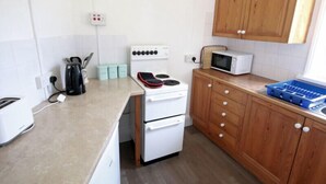 A compact kitchen with a white stove, a microwave, wooden cabinets, and a countertop with a toaster and storage containers. - Gullsway: Sandpiper (Wadebridge)