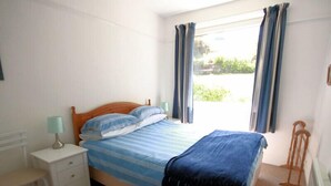A bedroom with a bed, a nightstand, a lamp, and a window with curtains. - Gullsway: Sandpiper (Wadebridge)