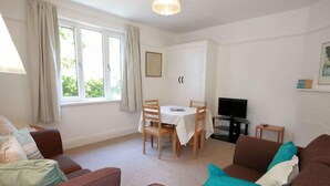A living room with a sofa, a dining table with chairs, a television, and a window with curtains. - Gullsway: Sandpiper (Wadebridge)