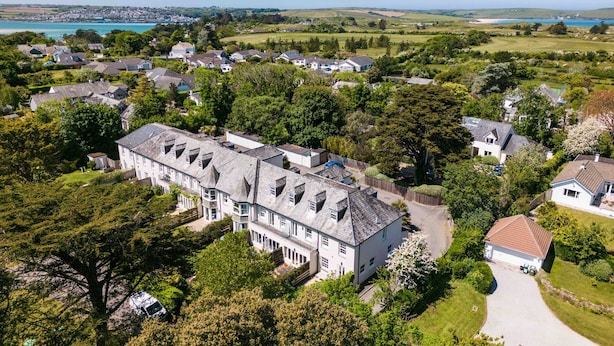 Een groot, meerlaags huis met een opvallend centraal gedeelte en een kleiner aanhangend deel, omringd door weelderige begroeiing en een uitzicht op de kust.
