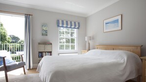 A bedroom with a large bed, a bedside table, a chair, a window with curtains, a bookshelf, and a painting on the wall. - Lowenna Manor 4 (Wadebridge)