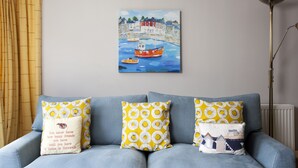 A living room with a blue sofa, yellow and white patterned cushions, a painting of boats on a blue background, and a floor lamp. - Lowenna Manor 4 (Wadebridge)