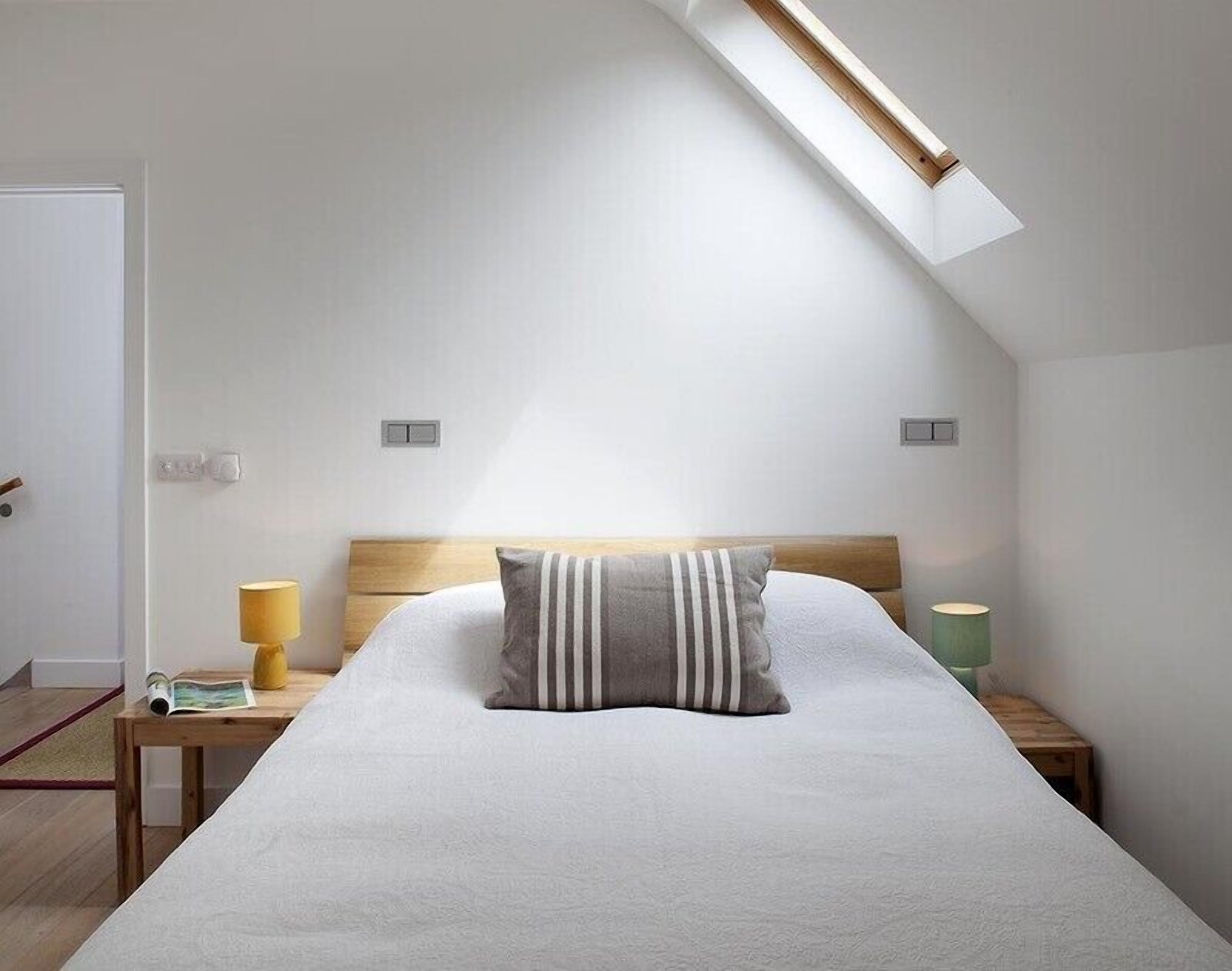 Un dormitorio con techo inclinado, una cama con almohada a rayas, dos mesitas de noche con lámparas y un tragaluz.