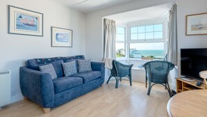 Una sala de estar con un sofá azul, dos sillas de mimbre, una mesa de madera, un televisor y una ventana con vista al mar.