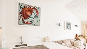 A modern kitchen with a large island, a sink, and a countertop. There is a framed artwork of a crab on the wall, a small cabinet, and a light fixture. - Slipway 10 (Wadebridge)