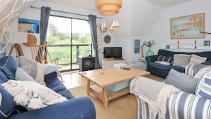 A cozy living room with a wooden coffee table, blue sofas, a TV, and a large window with curtains. - Rocklings (Wadebridge)